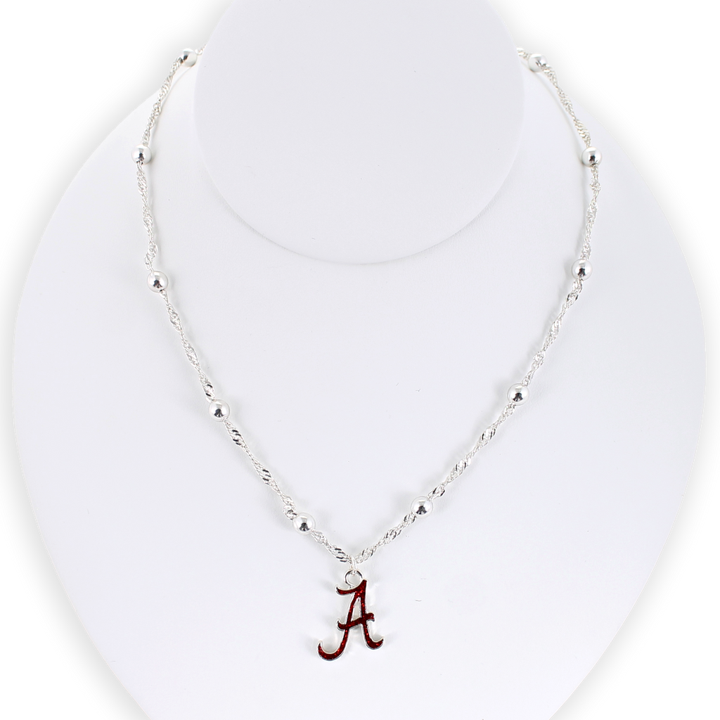 Show your school pride with the Alabama Glitter Enamel Logo Necklace by Seasons Jewelry. This silver beaded necklace features a crimson “A” logo pendant accented with sparkling glitter enamel, beautifully displayed on a white necklace stand—a shimmering tribute to University of Alabama spirit.