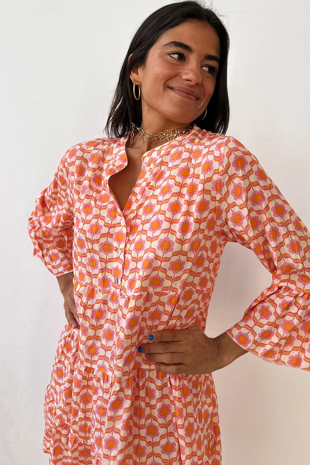 A person in a Shewin Orange Floral Button V Neck Bell Sleeve Mini Dress stands smiling with hands on hips, the boho floral print accentuating its V-neckline against a plain background.