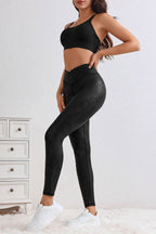 A woman stands indoors by a white dresser, styled in LA Society’s Black Crossed Dip Waist Sleek Leather Leggings, a black sports bra, and white sneakers.