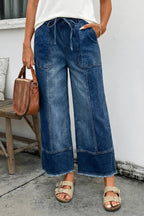 A person wearing 1 the Woman's Drawstring Seam Detail Raw Hem Wide Leg Denim Pants, paired with a white top and sandals, holds a brown handbag while standing beside a wooden bench against a white wall.
