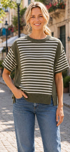 A woman stands outdoors on a cobblestone street, smiling at the camera with one hand in her pocket, wearing blue jeans and the Women’s Stripe Color Block Rib Knit Relaxed Top from Celeste Clothing—a casual green and white striped summer top crafted from rib knit fabric.