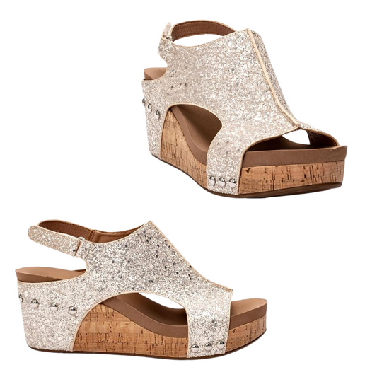 The Carley Wedge in Beige Glitter by Corky's is a pair of women's wedge sandals with cork soles, featuring glittery silver strap design and eye-catching metal stud details. These best-selling shoes from CORKY'S FOOTWEAR INC are perfect for adding a touch of sparkle to any outfit.