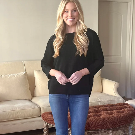 A blonde woman standing in a living room wearing jeans and a Carole Christian Black New Soft Sweater.