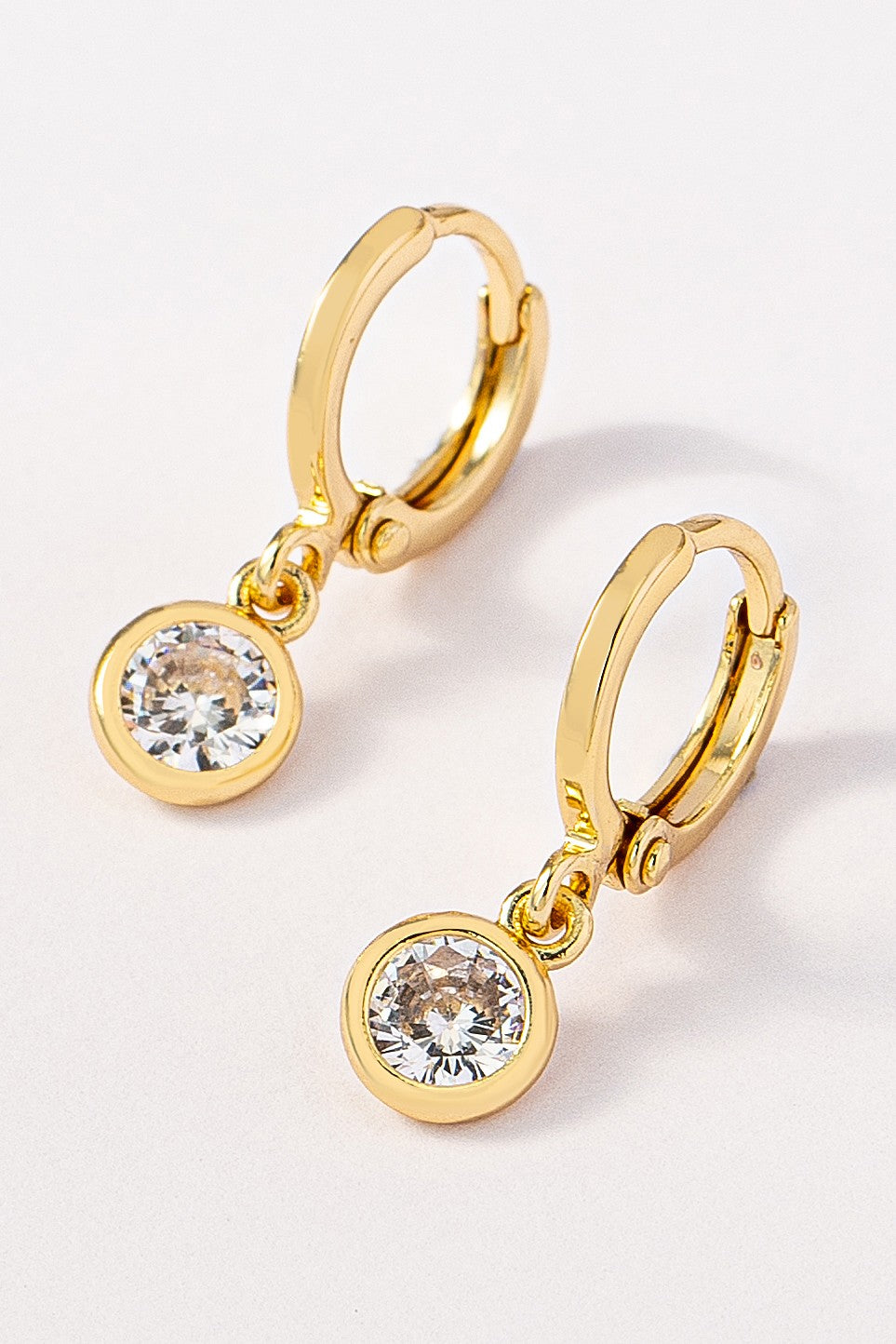 Pair of brass huggie hoops with a dangling round CZ pendant on a white background by FASHION GO. Main image
