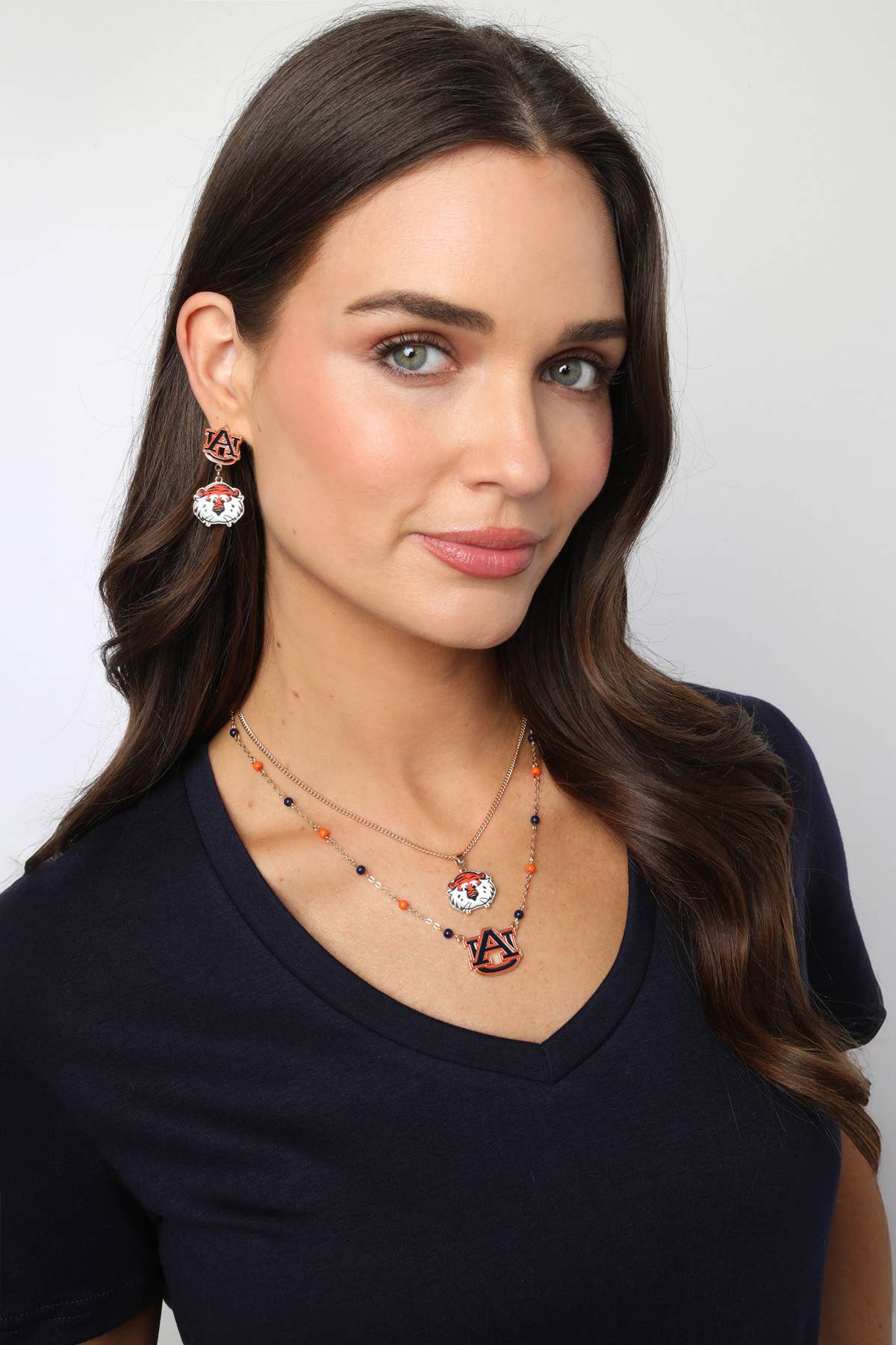 A woman with long brown hair poses against a plain white background wearing a navy V-neck shirt, orange and blue beaded necklaces, and Canvas Style’s Auburn Tigers Enamel Drop Earrings—the perfect game day jewelry.