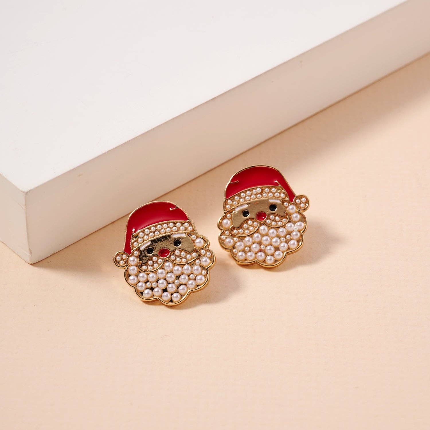 The Lilla Haven Pearl Santa Claus Stud Earrings, featuring charming red hats and beaded white beards, are displayed on a cream-colored surface beside a white rectangular object—making these Christmas Holiday Earrings the perfect seasonal accessory or thoughtful holiday gift. Main image