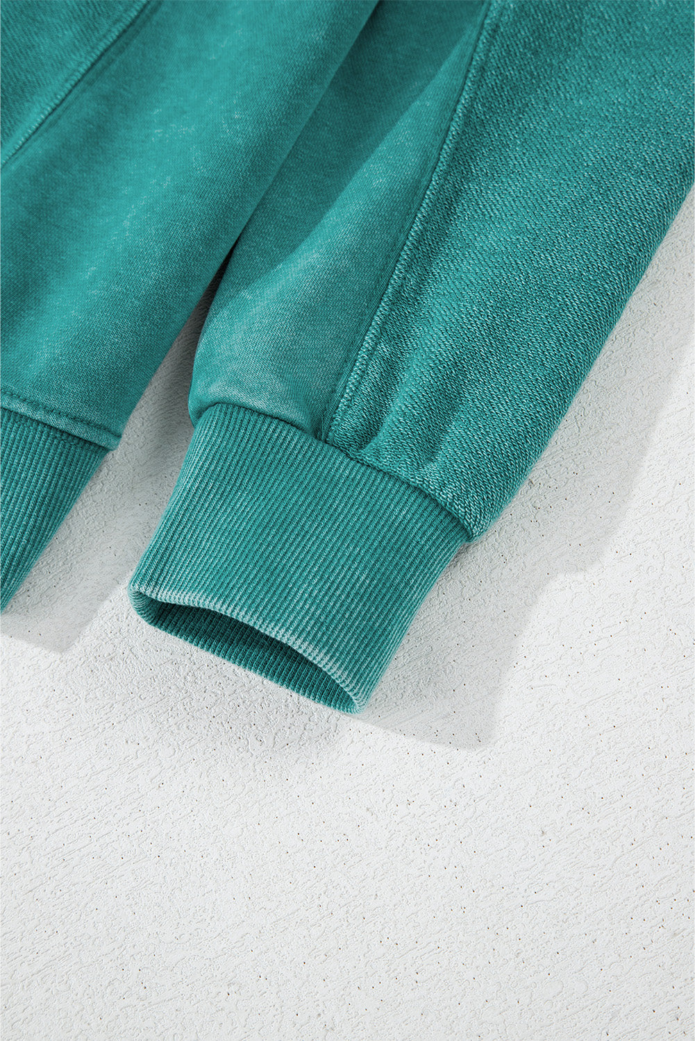 Close-up of a Notched Neck Exposed Seam Drop Shoulder Sweatshirt sleeve by Shewin, highlighting its high-quality teal fabric and ribbed cuff on a light surface.