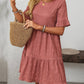 A woman wears the Suhkasana Loose Casual Short-Sleeve Dress in mauve, carrying a woven straw bag and accessorized with bracelets and a pendant necklace.