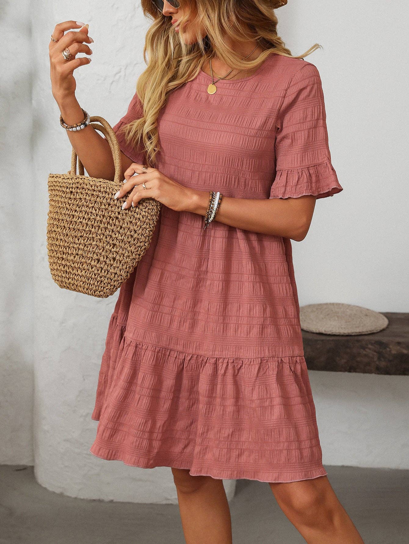 A woman wears the Suhkasana Loose Casual Short-Sleeve Dress in mauve, carrying a woven straw bag and accessorized with bracelets and a pendant necklace.