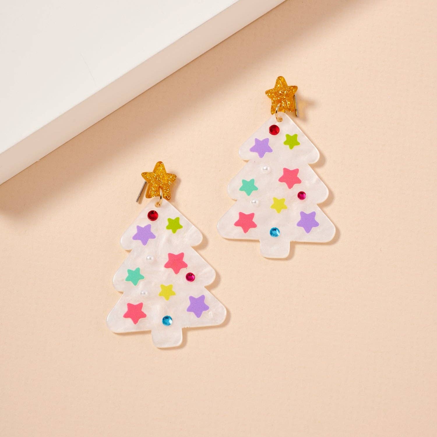 Add a festive touch to your holiday look with the Glittery Christmas Tree Drop Earrings by Lilla Haven. These holiday acrylic earrings feature two white Christmas tree shapes adorned with colorful star decorations and sparkling gold star tops, all shining beautifully on a beige background. Perfect as statement jewelry, these earrings are designed to make your celebrations bright and joyful while ensuring you stand out in style.