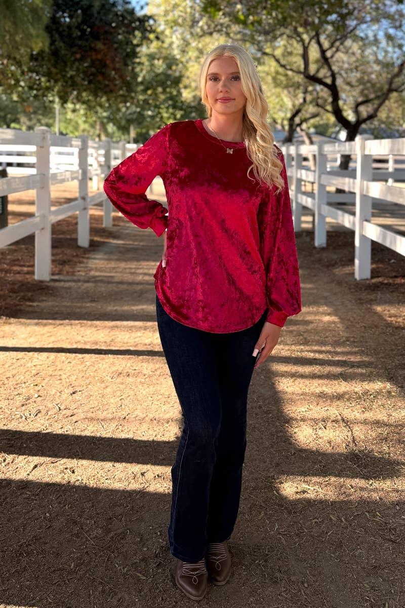 A woman stands outdoors by white fences, wearing the 24-Hrs Fashion Crushed Velvet Top with flowy sleeves, paired with dark jeans and brown loafers, trees in the background.