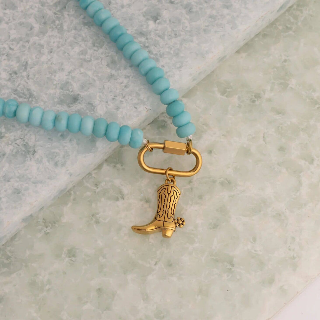 The WJW Stainless Steel Turquoise Chunky Bead Carabiner Necklace, featuring a gold cowboy boot charm, is displayed on a light textured surface.