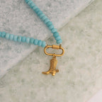 The WJW Stainless Steel Turquoise Chunky Bead Carabiner Necklace, featuring a gold cowboy boot charm, is displayed on a light textured surface.