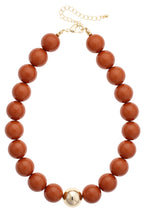 The Marjorie Game Day Beaded Necklace by Canvas Style features chunky, oversized round brown beads accented with a single central gold bead and is finished with a gold chain extender and clasp—an unforgettable statement necklace perfect for elevating any look.