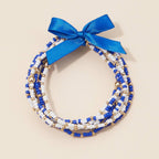 A stack of blue, white, and gold Game Day Seed Beaded Bracelet Set tied together with a blue ribbon bow.
