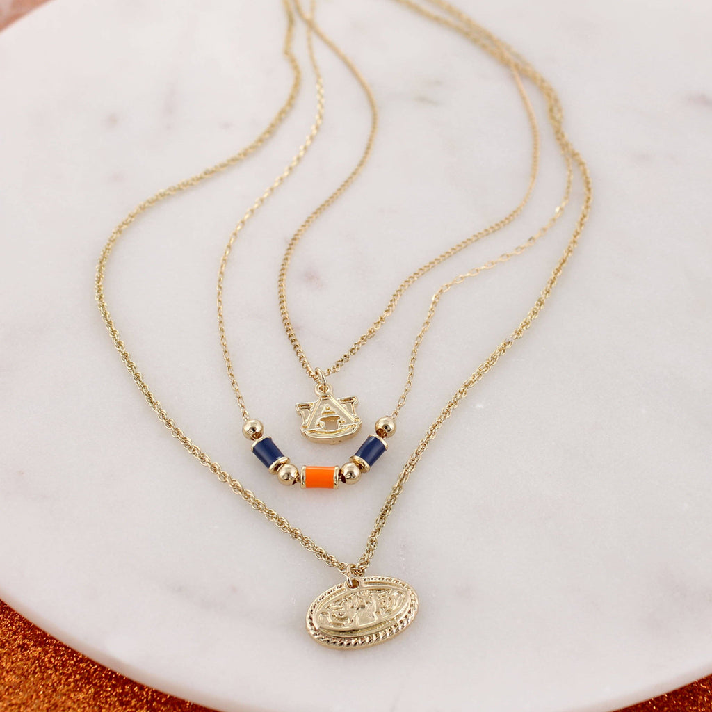 Showcase your Auburn spirit with the Auburn Gold & Tube Bead Trio Necklace Set by Seasons Jewelry. This elegant set features three gold layered necklaces displayed on a round marble surface—each adorned with unique pendants, including beaded accents and an emblem. Perfect for expressing your style or gifting a distinctive piece of War Eagle jewelry, this necklace set beautifully combines timeless design with Auburn University pride.