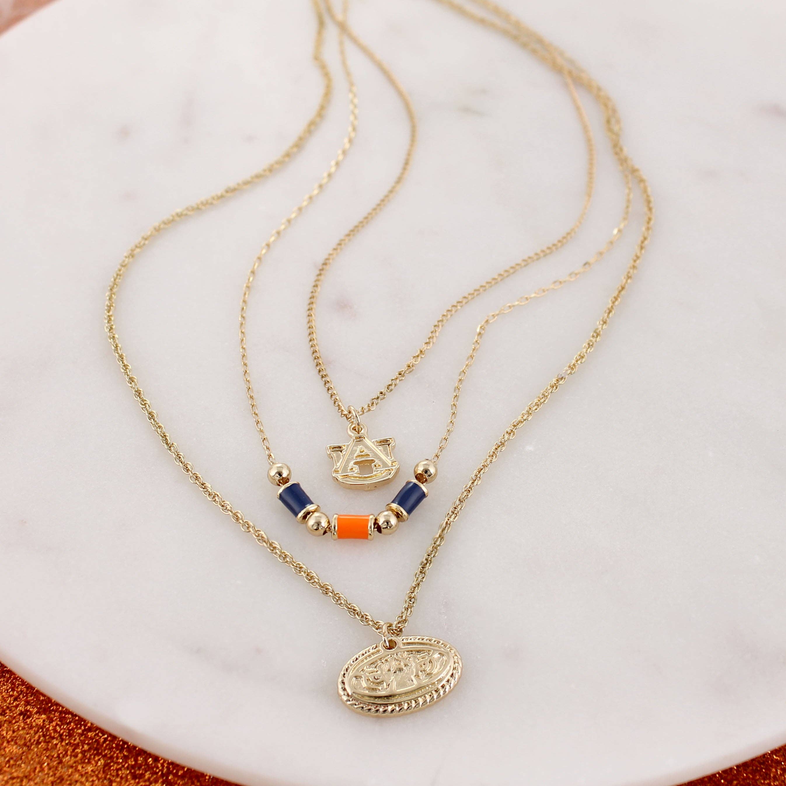 Showcase your Auburn spirit with the Auburn Gold & Tube Bead Trio Necklace Set by Seasons Jewelry. This elegant set features three gold layered necklaces displayed on a round marble surface—each adorned with unique pendants, including beaded accents and an emblem. Perfect for expressing your style or gifting a distinctive piece of War Eagle jewelry, this necklace set beautifully combines timeless design with Auburn University pride.