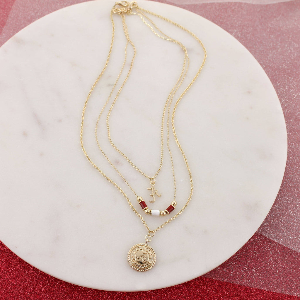 Three gold necklaces from the Alabama Gold & Tube Bead Trio Necklace Set by Seasons Jewelry displayed on a round marble surface: one with a University of Alabama medallion, one embellished with red and white tube beads reminiscent of Bama jewelry, and one featuring a small letter "J" charm.