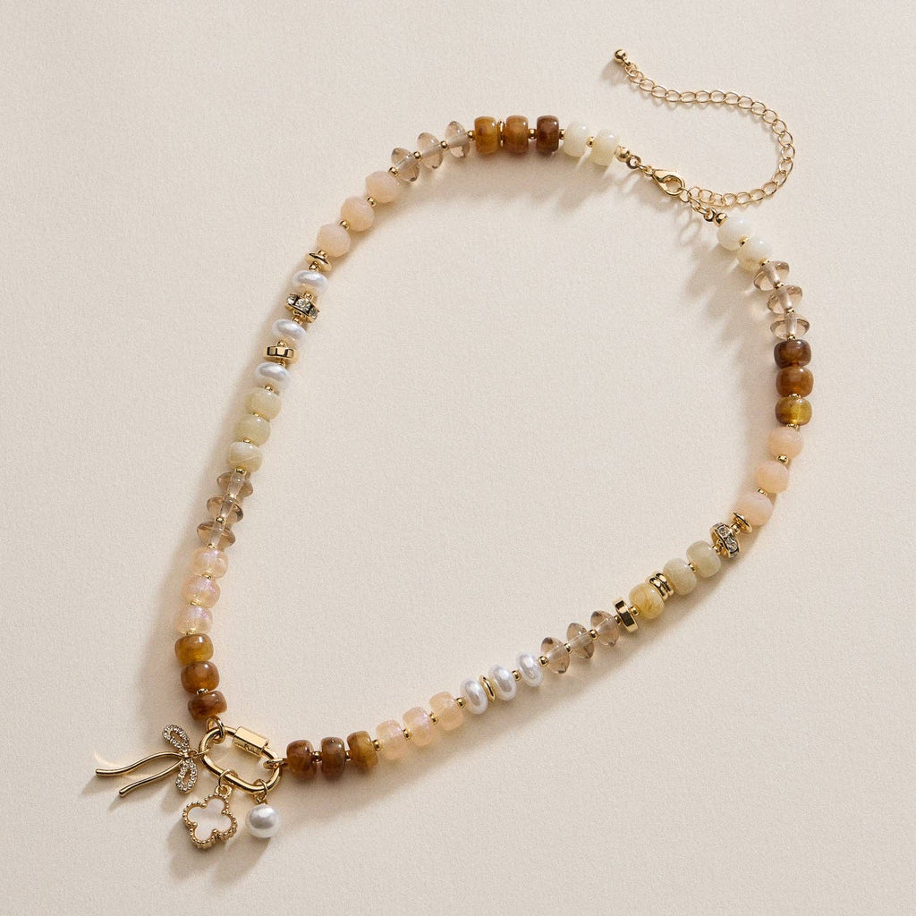Lilla Haven’s Textured Glass Bead Necklace with Charm Trio features mixed neutral-toned beads, gold-toned spacers, metallic accents, and several gold and pearl charms, displayed on a cream background.