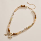 Lilla Haven’s Textured Glass Bead Necklace with Charm Trio features mixed neutral-toned beads, gold-toned spacers, metallic accents, and several gold and pearl charms, displayed on a cream background.