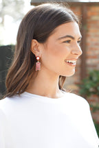 A woman with straight brown hair, dressed in a white top and smiling as she looks to her left outdoors against a blurred brick wall, adds a bold touch to her look with the Canvas Style Alabama Crimson Tide Resin Block Letter Earrings in Crimson—an NCAA officially licensed accessory.