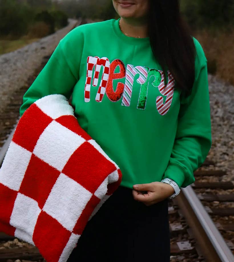 Person wearing a Faire Green Merry Sequin Christmas Sweatshirt with the word “Merry” in sequined, candy-cane-striped letters, holding a red and white checkered blanket.