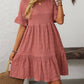 A woman with long wavy blonde hair wears the Suhkasana Loose Casual Short-Sleeve Dress in dusty pink, styled with a woven bag, bracelets, and a necklace.