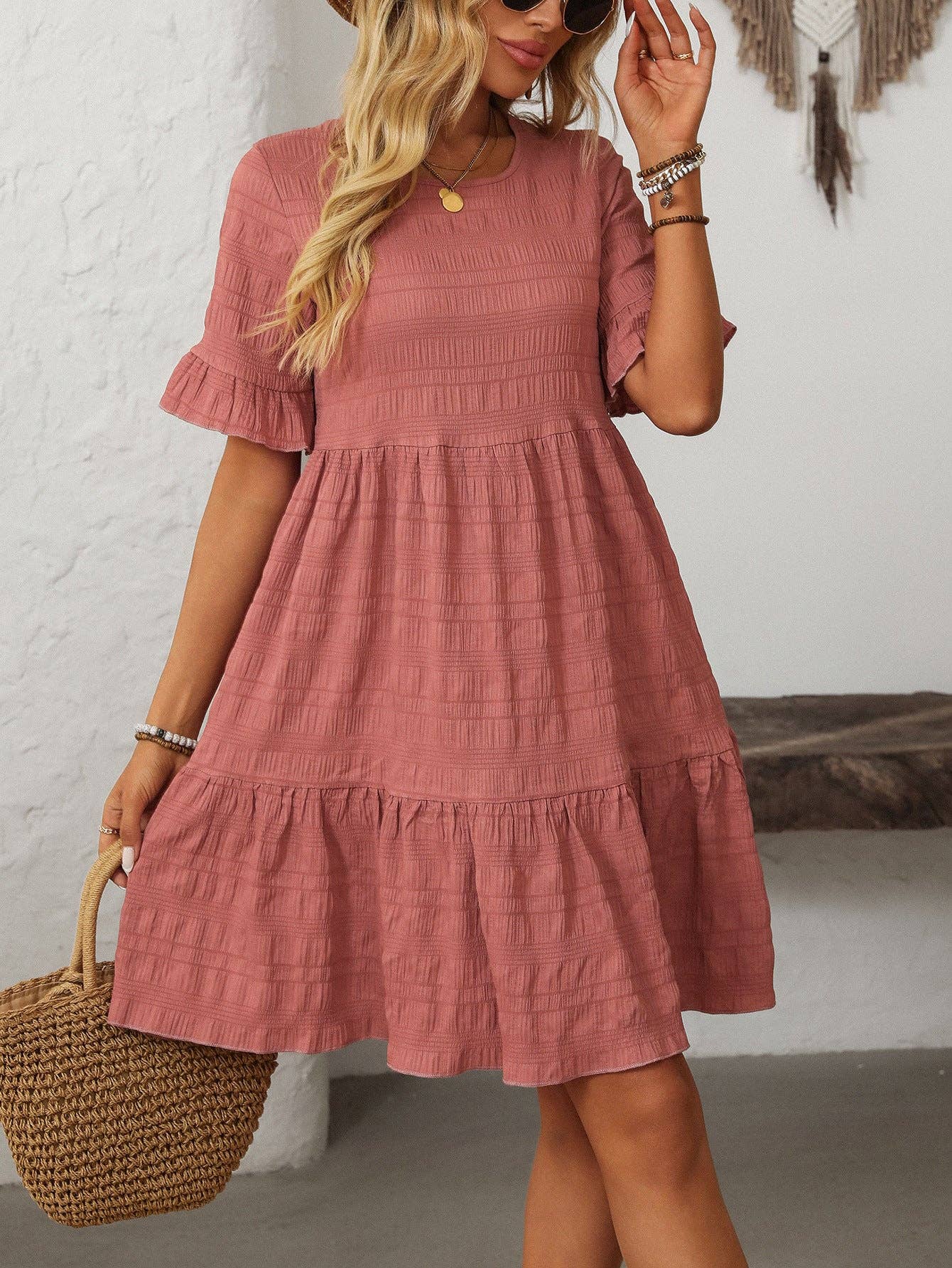 A woman with long wavy blonde hair wears the Suhkasana Loose Casual Short-Sleeve Dress in dusty pink, styled with a woven bag, bracelets, and a necklace.