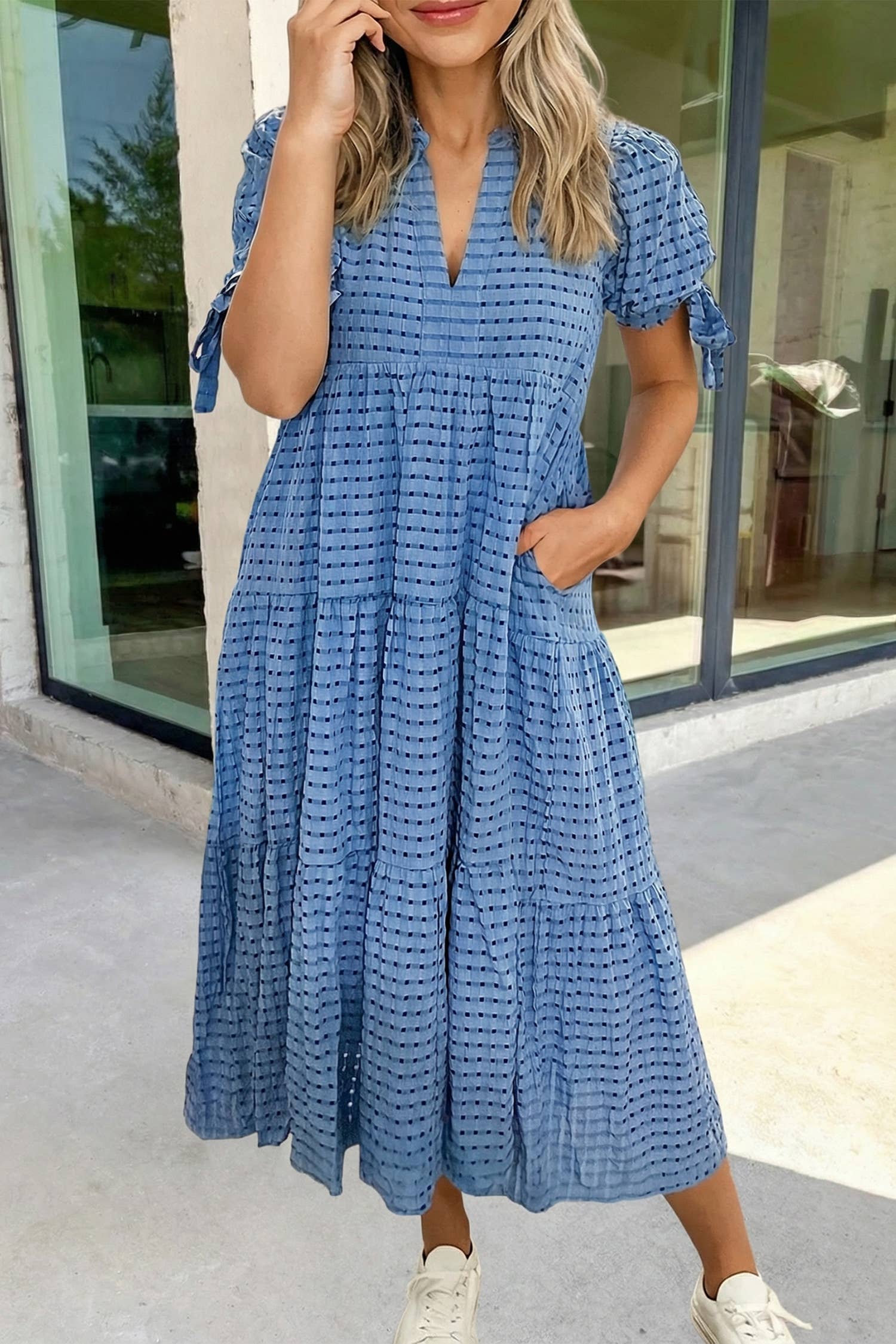 A woman stands outside by glass doors, one hand in her pocket, wearing the Aapparella Textured Gingham Bow Tie Tiered Midi Dress (S-XL) with short sleeves and white sneakers.