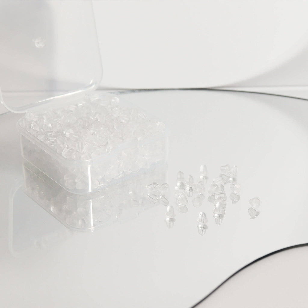 A clear plastic container from WJW holds one pair of their Silicone Earring Backs, with several secure, transparent earring backs scattered on a reflective surface nearby.