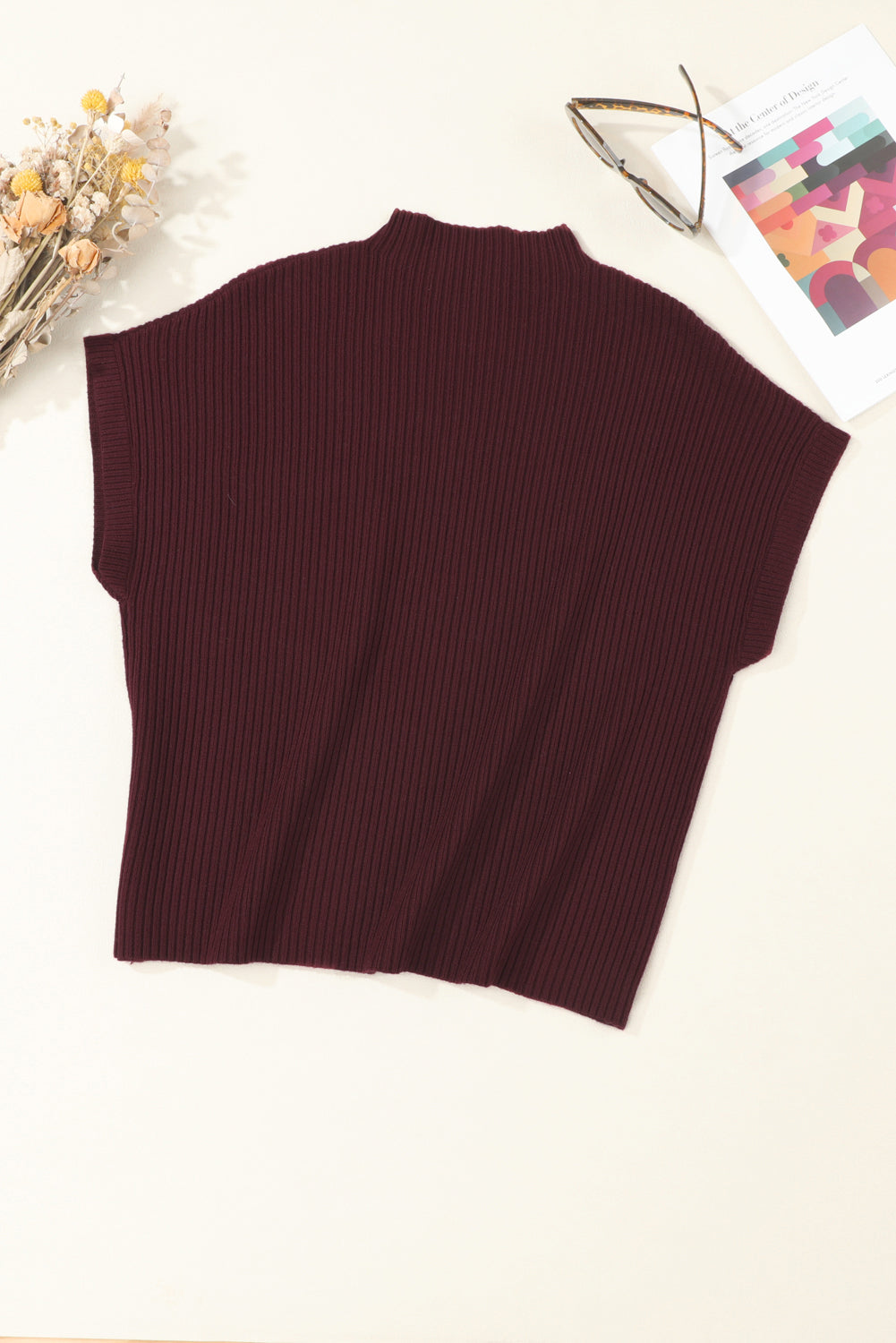 The Shewin Patch Pocket Ribbed Knit Short Sleeve Sweater in dark red is laid flat beside a dried flower bouquet. Ideal for modern casual looks and transitional weather, it pairs well with glasses and a colorful abstract notebook.