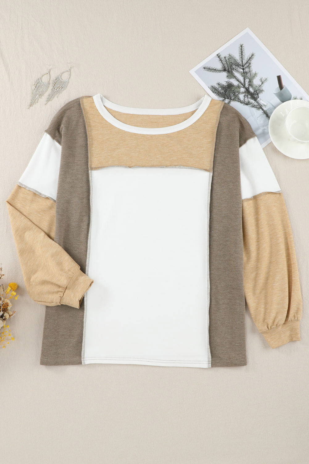 The Khaki Color Block Exposed Seam Long Sleeve Top by Shewin is laid flat, showcasing its beige, white, and gray tones. Beside the shirt, a white teacup and saucer, a photo with pine branches, and decorative earrings add to the aesthetic. This online exclusive piece features a stylish patchwork design.