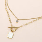 Sensibling Corp.’s Stainless Steel Mother of Pearl Rectangular Frame Toggle Pendant Round Stone Accent Layered Necklace features two elegant layers: one with a small round stone accent pendant and the other with a toggle clasp and a rectangular mother of pearl charm, beautifully displayed on a light surface.