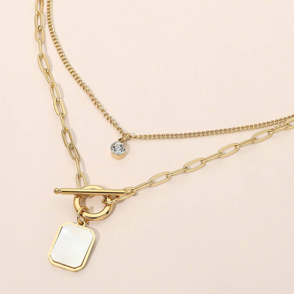 Sensibling Corp.’s Stainless Steel Mother of Pearl Rectangular Frame Toggle Pendant Round Stone Accent Layered Necklace features two elegant layers: one with a small round stone accent pendant and the other with a toggle clasp and a rectangular mother of pearl charm, beautifully displayed on a light surface.