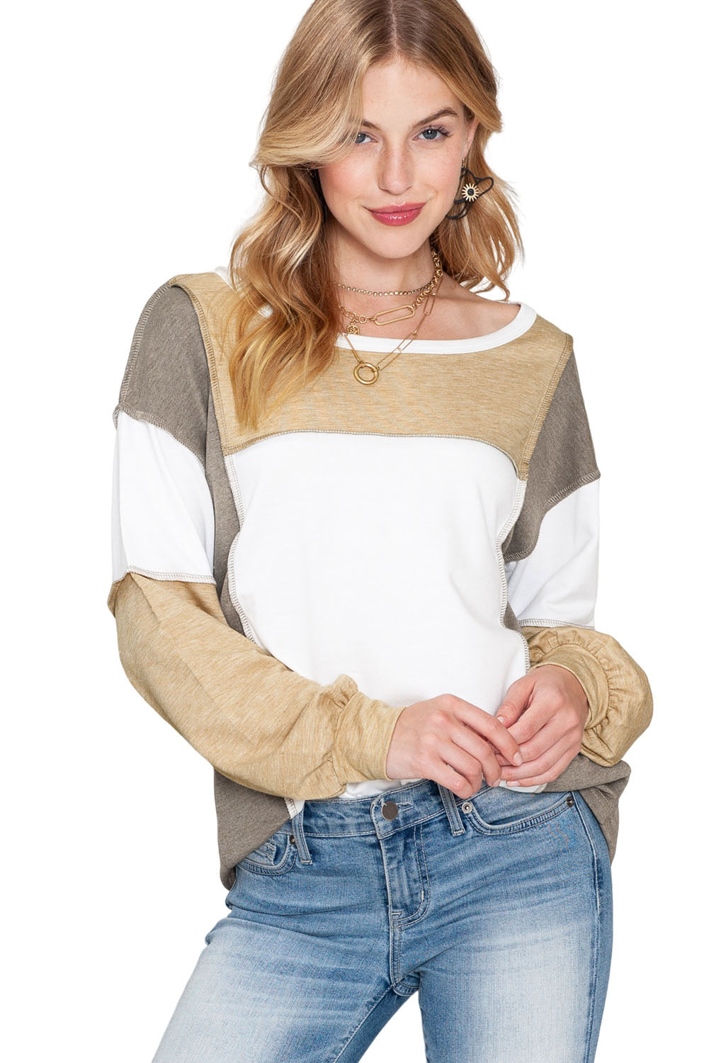 A woman with blonde hair is wearing the Shewin Khaki Color Block Exposed Seam Long Sleeve Top, featuring a unique patchwork design in shades of beige, white, and gray. She has paired it with light blue jeans and is standing while looking at the camera with a slight smile.