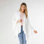 A woman stands against a plain background wearing the COCO + CARMEN Kiara Lightweight Wrap in white over a light pink top and blue jeans. This stylish, one-size-fits-most piece is perfect for effortless layering.
