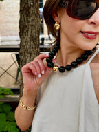 A woman standing outdoors near a tree wears the Marjorie Game Day Beaded Necklace by Canvas Style—a chunky beaded necklace with a gold accent—paired with gold earrings, a gold bracelet, sunglasses, and a white sleeveless top.
