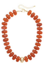 The San Sebastián Beaded Resin Necklace by Canvas Style features chunky orange resin beads with a single large gold bead at the center and a gold adjustable chain clasp. This statement necklace adds a vibrant touch to any look.