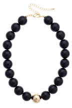 Introducing the Marjorie Game Day Beaded Necklace by Canvas Style—this statement piece features oversized black beads accented with a single gold bead at the center and is finished with an adjustable gold chain clasp. Perfect for adding bold style to any outfit!.