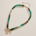Introducing the Lilla Haven Textured Glass Bead Necklace with Charm Trio, featuring vibrant green and gold glass beads, lustrous pearl accents, and three distinctive gold charms—an eye, a hand, and a horseshoe—each delicately separated by gold-toned spacers set against a beige background. This elegant charm necklace from Lilla Haven seamlessly blends refined style with meaningful symbolism.
