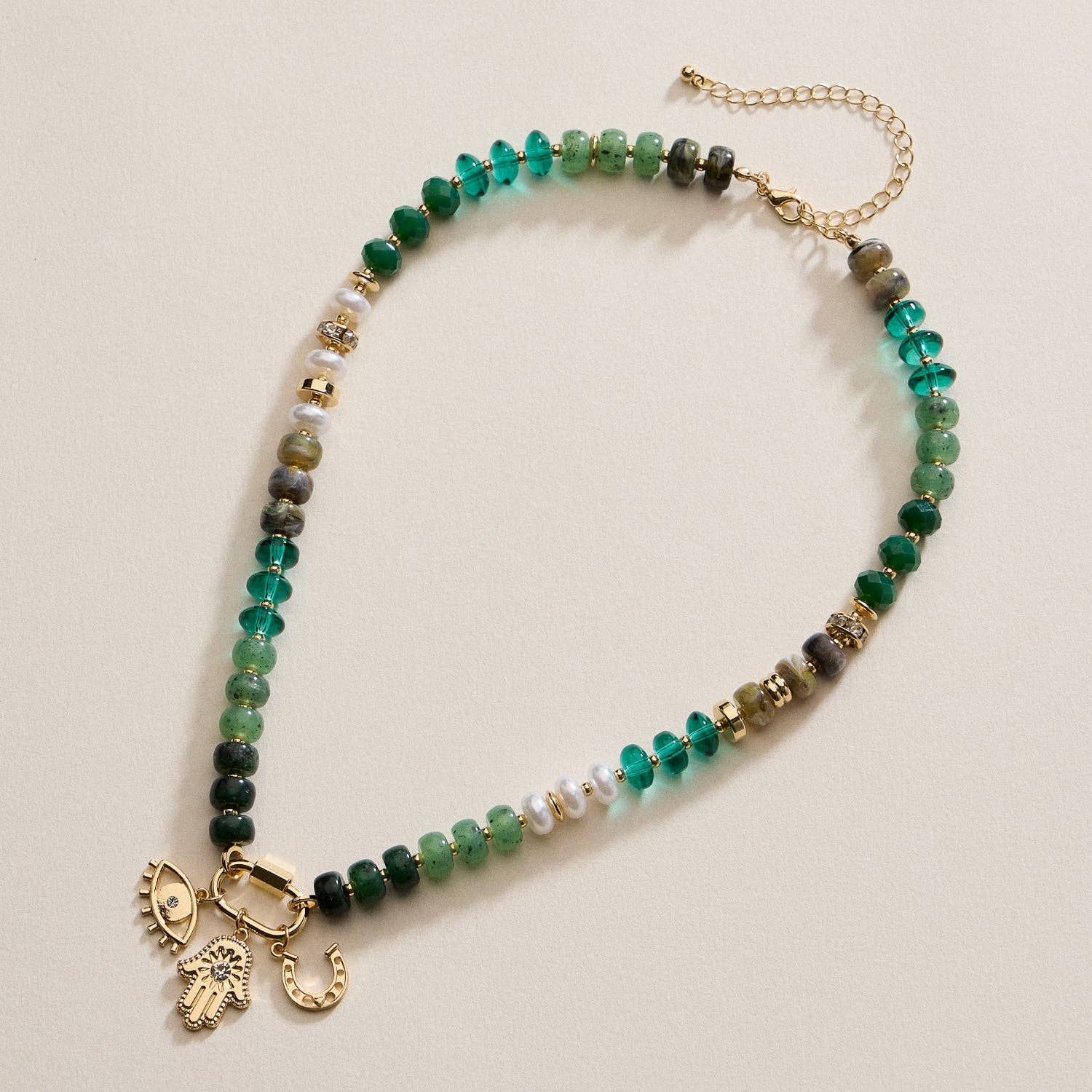 Introducing the Lilla Haven Textured Glass Bead Necklace with Charm Trio, featuring vibrant green and gold glass beads, lustrous pearl accents, and three distinctive gold charms—an eye, a hand, and a horseshoe—each delicately separated by gold-toned spacers set against a beige background. This elegant charm necklace from Lilla Haven seamlessly blends refined style with meaningful symbolism.