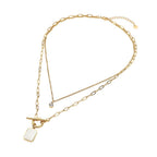 Introducing the Stainless Steel Mother of Pearl Rectangular Frame Toggle Pendant Round Stone Accent Layered Necklace by Sensibling Corp.—a gold double-layered necklace featuring mixed chain styles, with one layer adorned by a small round stone accent and the other showcasing a rectangular mother of pearl frame pendant with a toggle clasp. Crafted from durable stainless steel for lasting style and modern elegance.