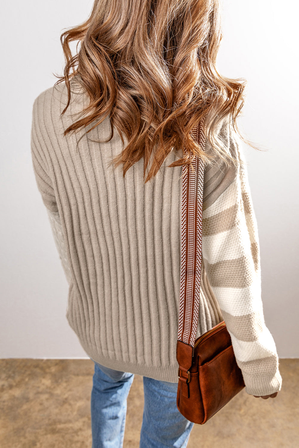 A person with wavy hair wears a Shewin Khaki Cable Knit Colorblock Crew Neck Drop Shoulder Sweater, pairs it with jeans, and carries a brown crossbody bag.