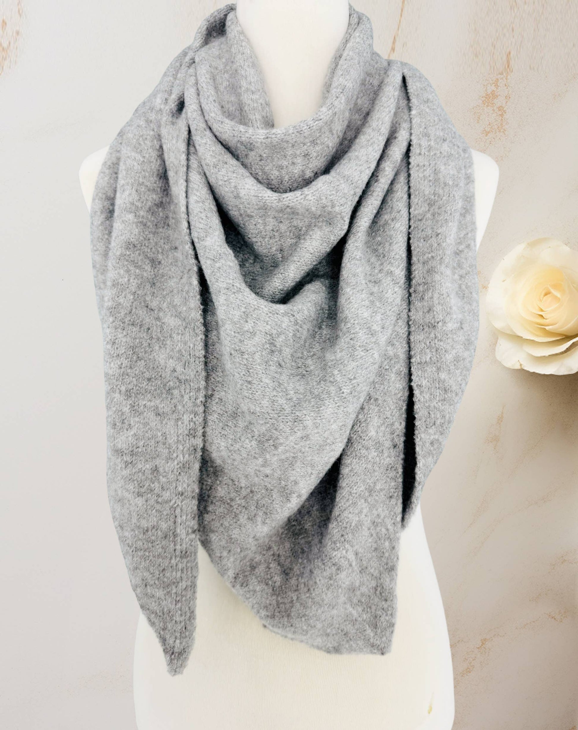 A JC Sunny Fashion Cashmere-Blend Knit Triangle Shawl with Cross-Front Design in light gray is draped around a white mannequin bust, with a cream-colored flower in the background.