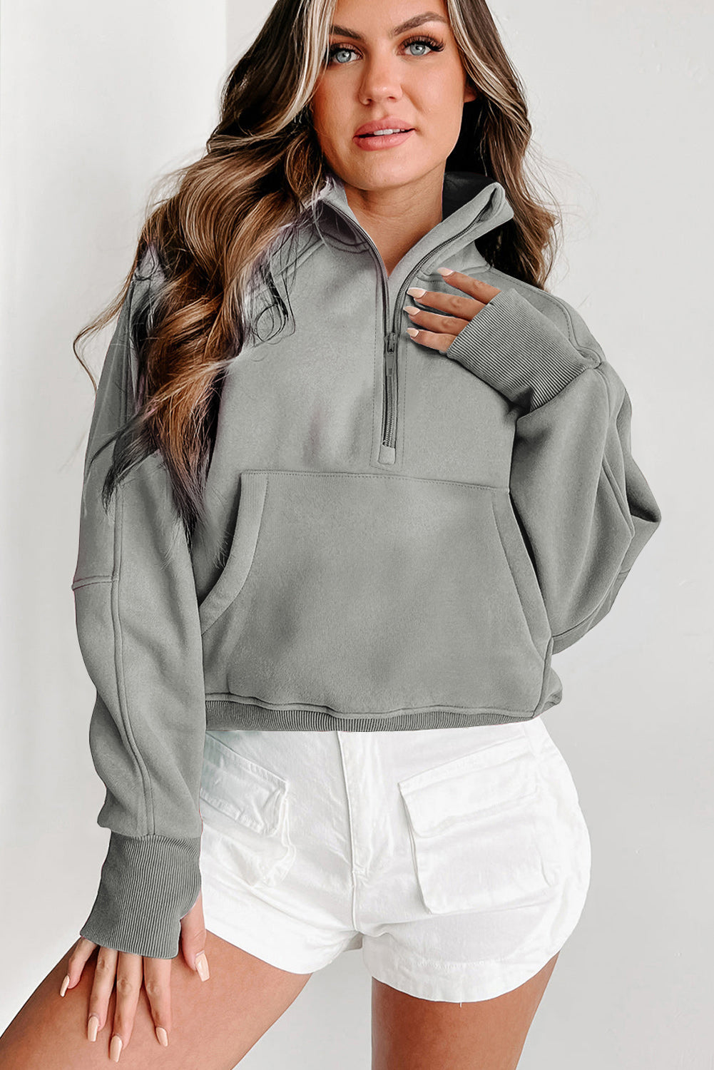 A woman with long, wavy hair wears a versatile Shewin Gray Zip Up Stand Collar Ribbed Thumbhole Sleeve Sweatshirt and white shorts, standing with her back partially turned.