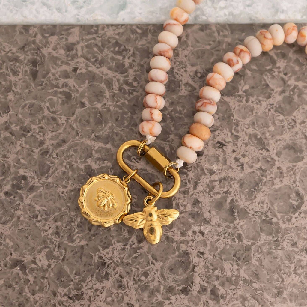 WJW Stainless Steel Chunky Glass Bead Carabiner Necklace with cream and tan stones, accented by gold bee and medallion charms on a gold clasp, displayed on a gray textured surface.