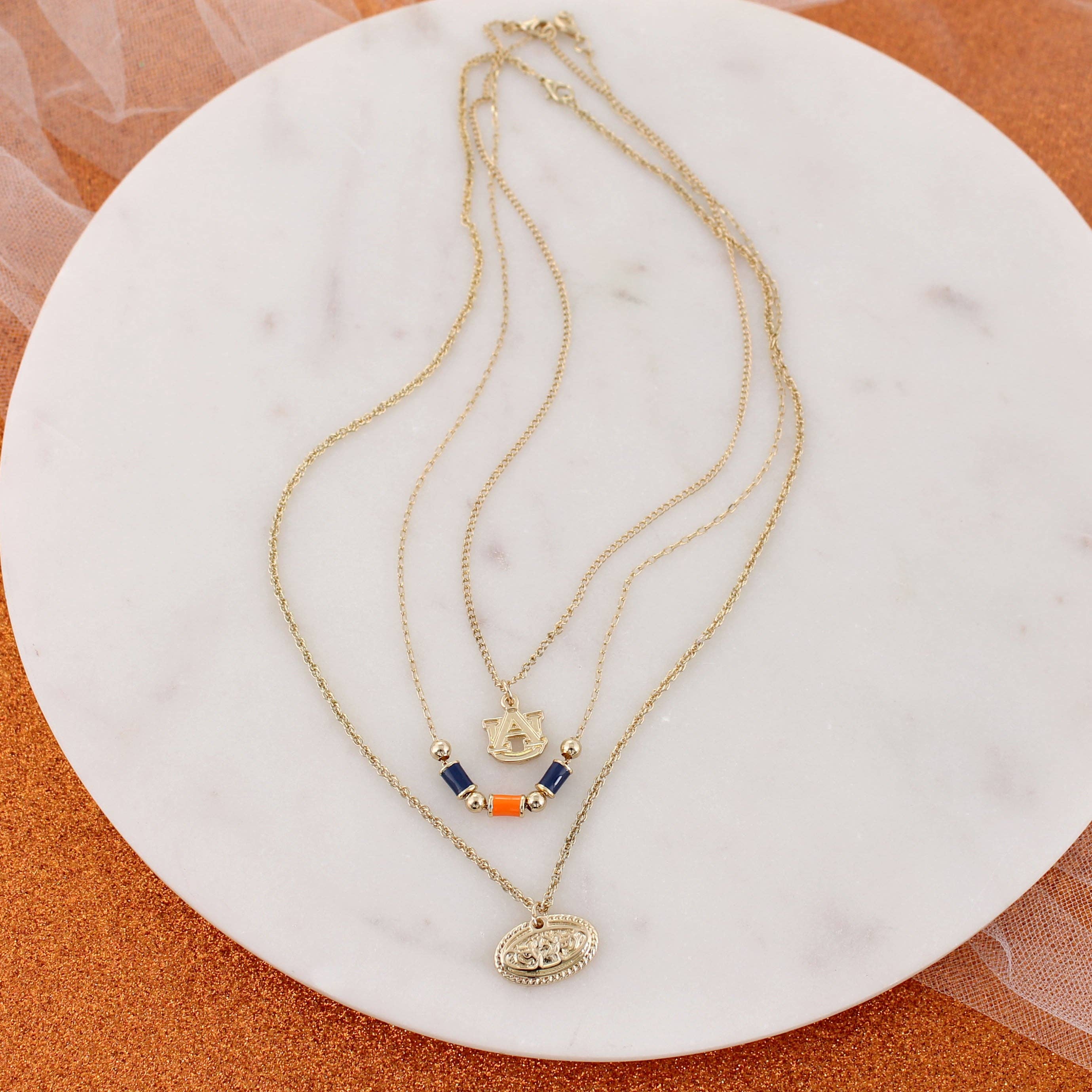 The Auburn Gold & Tube Bead Trio Necklace Set by Seasons Jewelry features three gold necklaces with unique pendants—including colored beads, a shield charm, and an oval pendant—artfully displayed on a round white marble surface, making it perfect for stylish layering or showing off your Auburn University spirit.