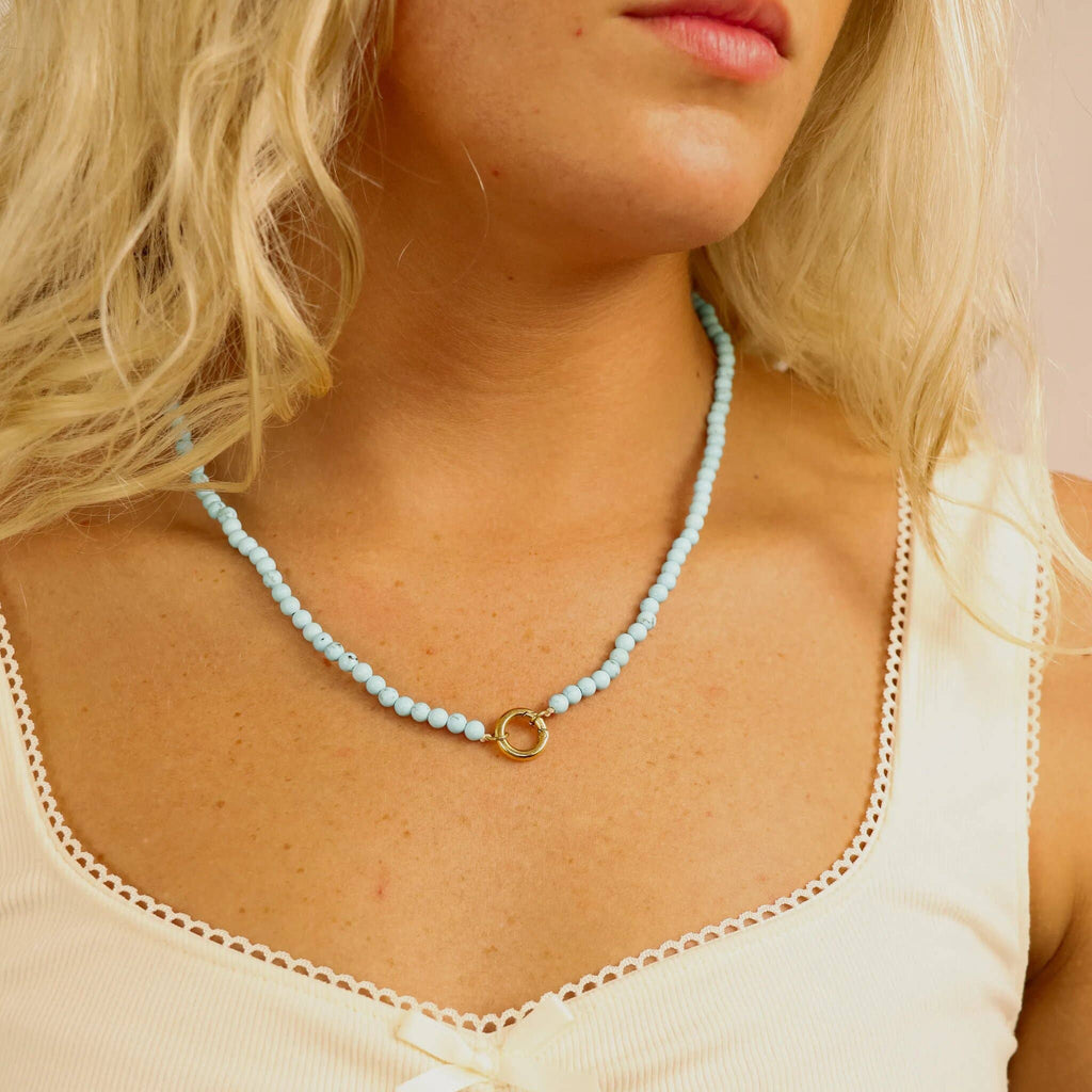 A woman wearing the WJW Semi Precious Natural Stone Charm Keeper Necklace, featuring a light blue semi precious stone strand and a circular 18K Gold PVD coated clasp, styled elegantly over a cream tank top.