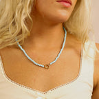 A woman wearing the WJW Semi Precious Natural Stone Charm Keeper Necklace, featuring a light blue semi precious stone strand and a circular 18K Gold PVD coated clasp, styled elegantly over a cream tank top.