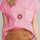 A person wearing a Shewin Patch Pocket Ribbed Knit Short Sleeve Sweater in pink, accented by a brown pendant necklace and paired with a brown button-up skirt.